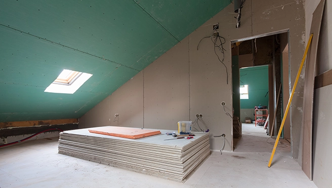 A partially finished attic renovation featuring green drywall, skylight, stacked drywall sheets, and scattered tools.