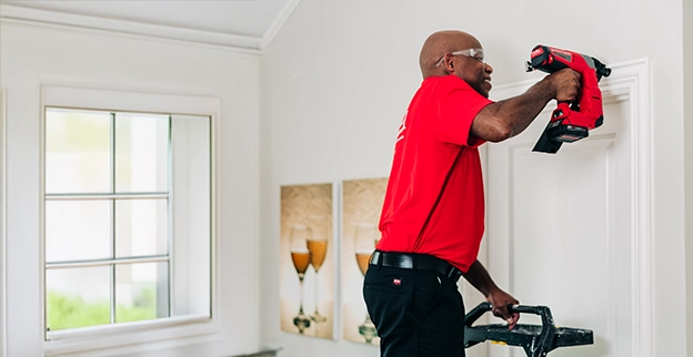 A person in a red shirt operates a nail gun while standing on a ladder.