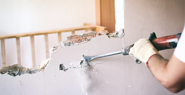 A construction worker wearing gloves uses a jackhammer to break through a wall, creating dust and debris.