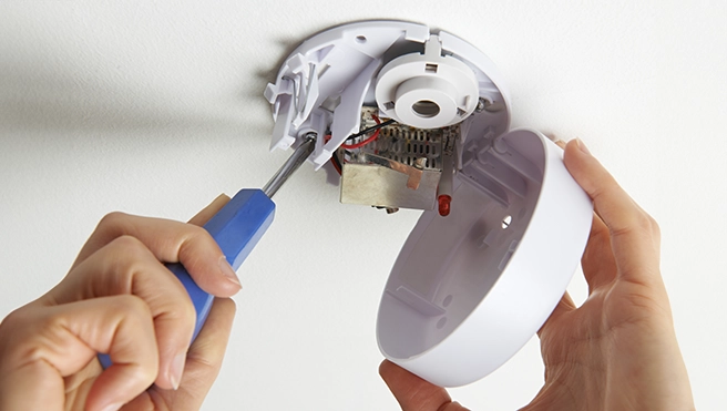 A person uses a screwdriver to install a white smoke detector on a ceiling.