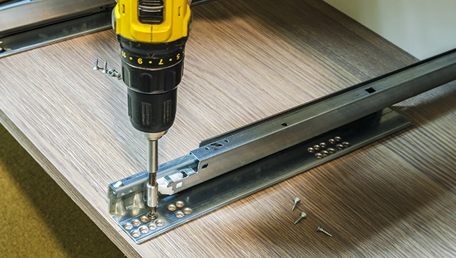 A power drill secures metal drawer slides to a wooden surface.