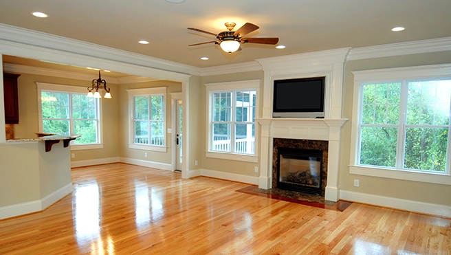 Bright living room with hardwood floors, a ceiling fan, fireplace, and large windows showcasing a green view outside.