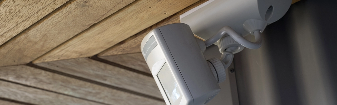 A white security camera mounted on a wooden ceiling, with its lens pointing downwards and the surrounding textures visible.
