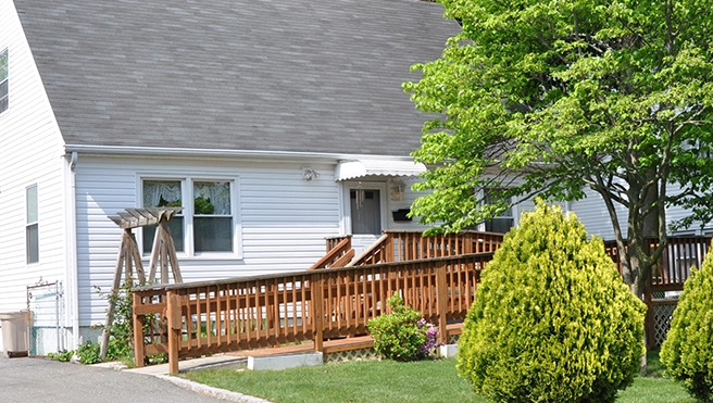 A cozy white house with a wooden ramp leading to the front door, surrounded by green shrubs and a large tree.