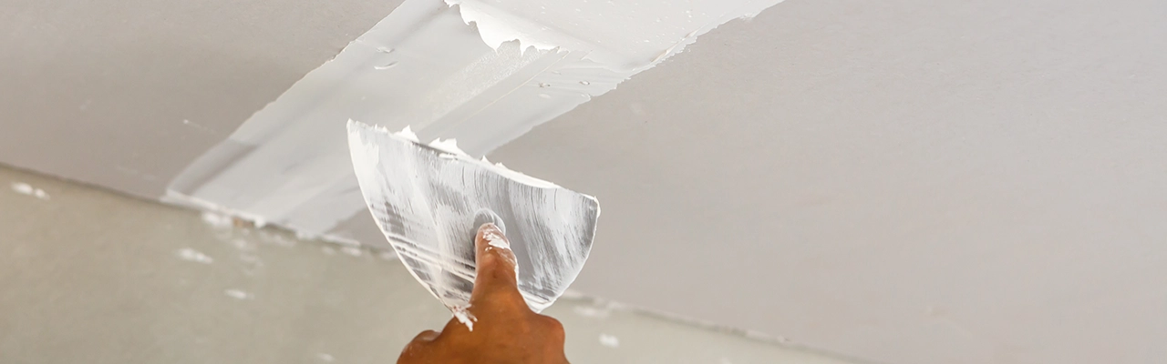 A person's hand holds a putty knife, smoothing white joint compound on a ceiling, preparing it for painting or finishing.