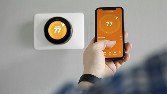 A hand holds a smartphone displaying a thermostat app with a wall-mounted smart thermostat showing 77°F.