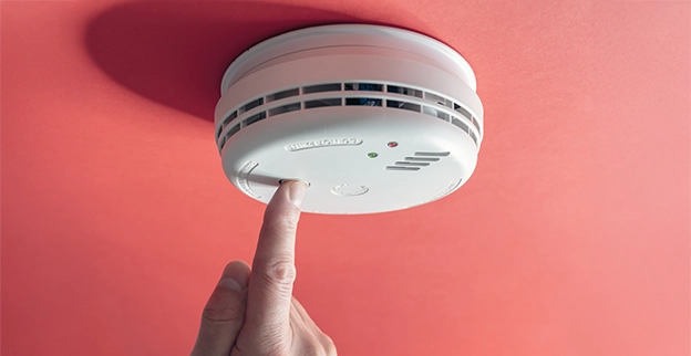 A hand presses the test button on a smoke detector mounted on a red ceiling.