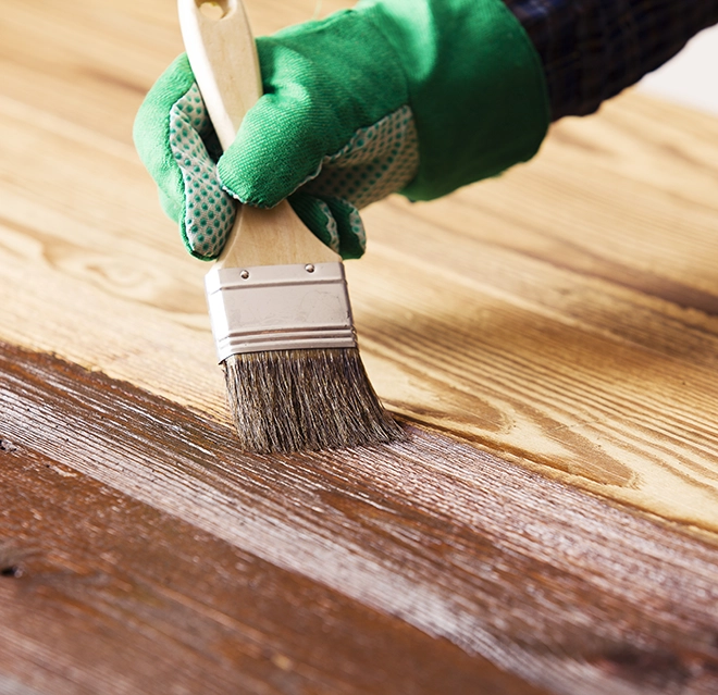 A gloved hand, holding a brush, applies varnish to a wooden surface.