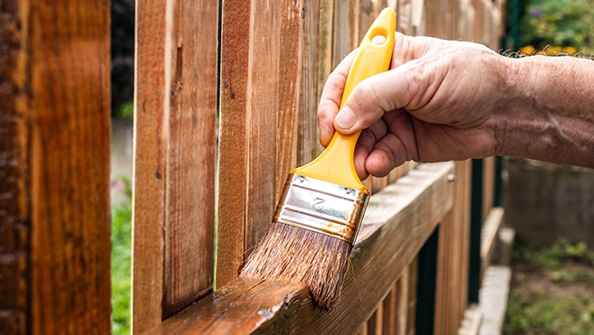A hand holds a yellow paintbrush, applying varnish to a wooden fence.