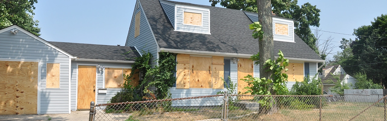 A boarded-up house with plywood covering windows and doors, surrounded by overgrown vegetation and a chain-link fence.