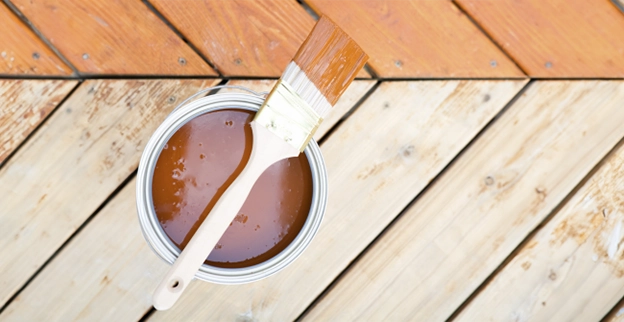 A paint can filled with brown stain and a brush rests on a wooden deck.