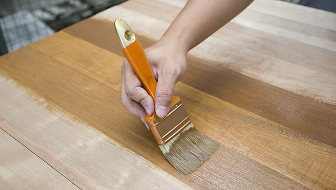 A hand holding a brush applies wood finish to a wooden surface.