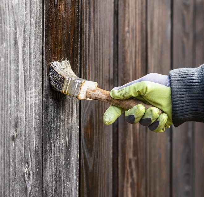 A gloved hand paints a wooden fence with a brush.