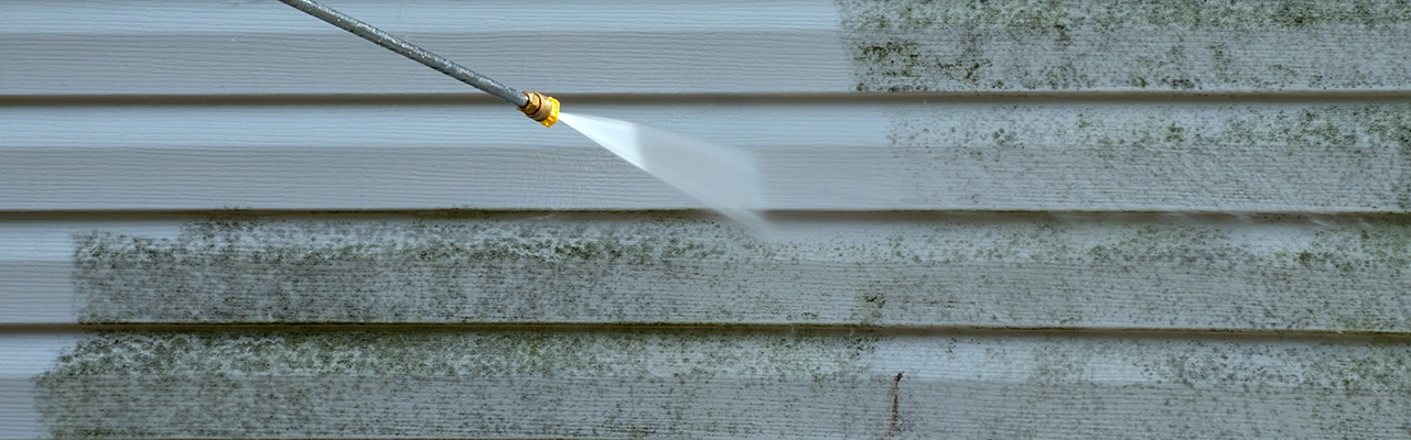 A power washer nozzle sprays water on a dirty, moldy exterior wall, cleaning away grime and revealing a fresh, white surface.