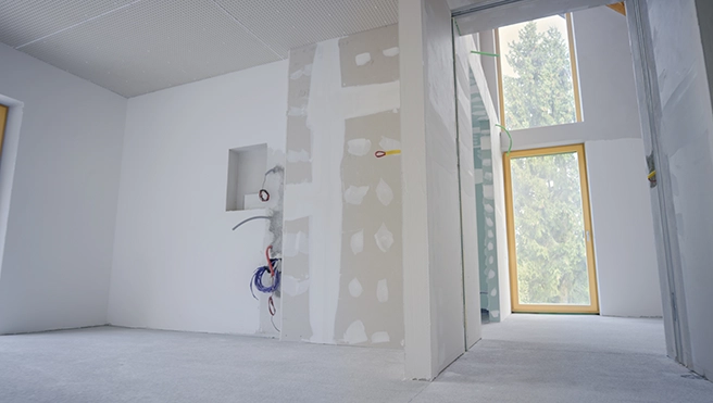 Interior of a partially finished room with drywall and construction supplies, featuring large windows and unfinished walls.