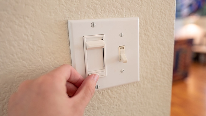 A hand reaches to toggle a white light switch on a textured wall.