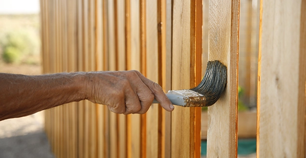 A close-up of a hand painting a wooden fence with a brush.