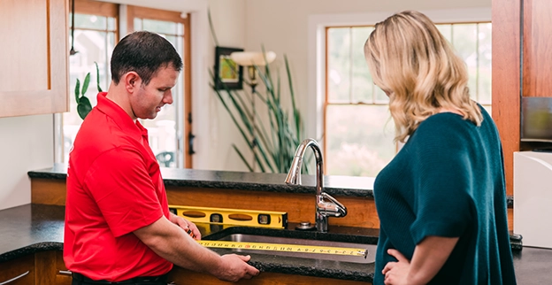 A person measures a countertop, while a woman observes attentively in a bright kitchen.
