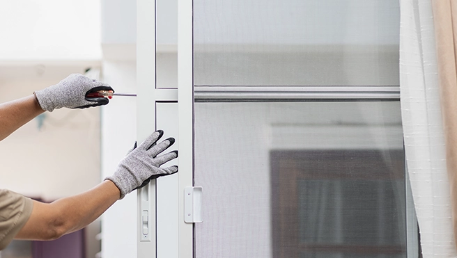 A person wearing gray gloves is adjusting a white framed window screen with a small tool in hand.