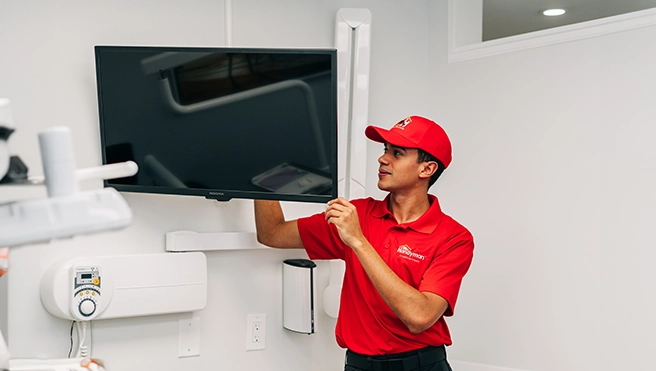 A technician in a red uniform installs a flat-screen TV on a white wall.