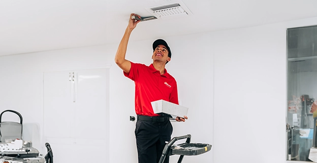 A worker in a red shirt uses a tool to adjust a ceiling vent in a clean, modern room with minimal furnishings.
