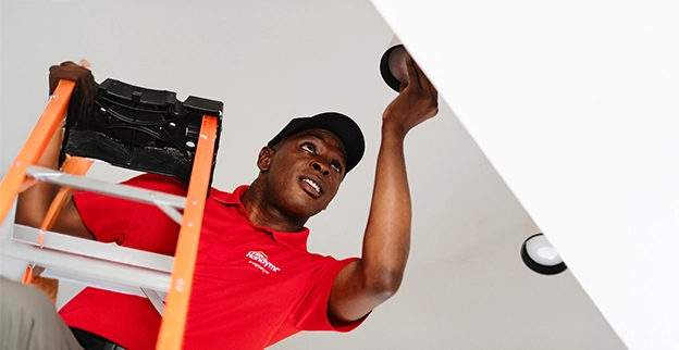 Handyman stands on a ladder, installing recessed lighting fixtures on a white ceiling.