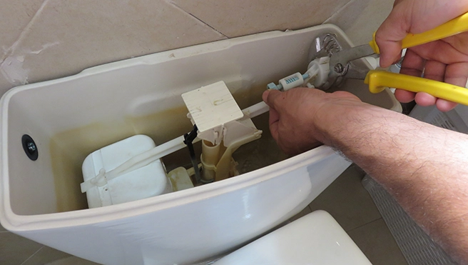 A person repairs a toilet tank, using pliers on the internal components.