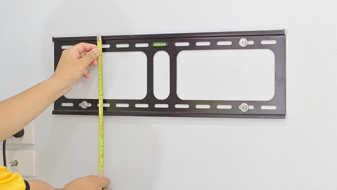 A person measures distance a mounted TV wall bracket on a light-colored wall.