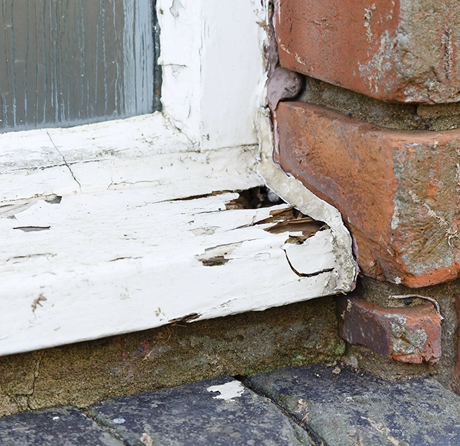 A weathered wooden window sill with paint peeling and cracks, adjacent to rough brickwork and cobblestone.