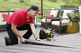 A worker on a wooden deck, using a saw to cut a plank, with a toolbox and a yellow drill nearby.