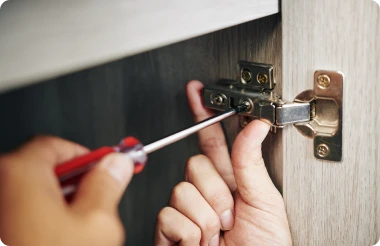 A person securing a metal hinge on a cabinet door with a screwdriver.