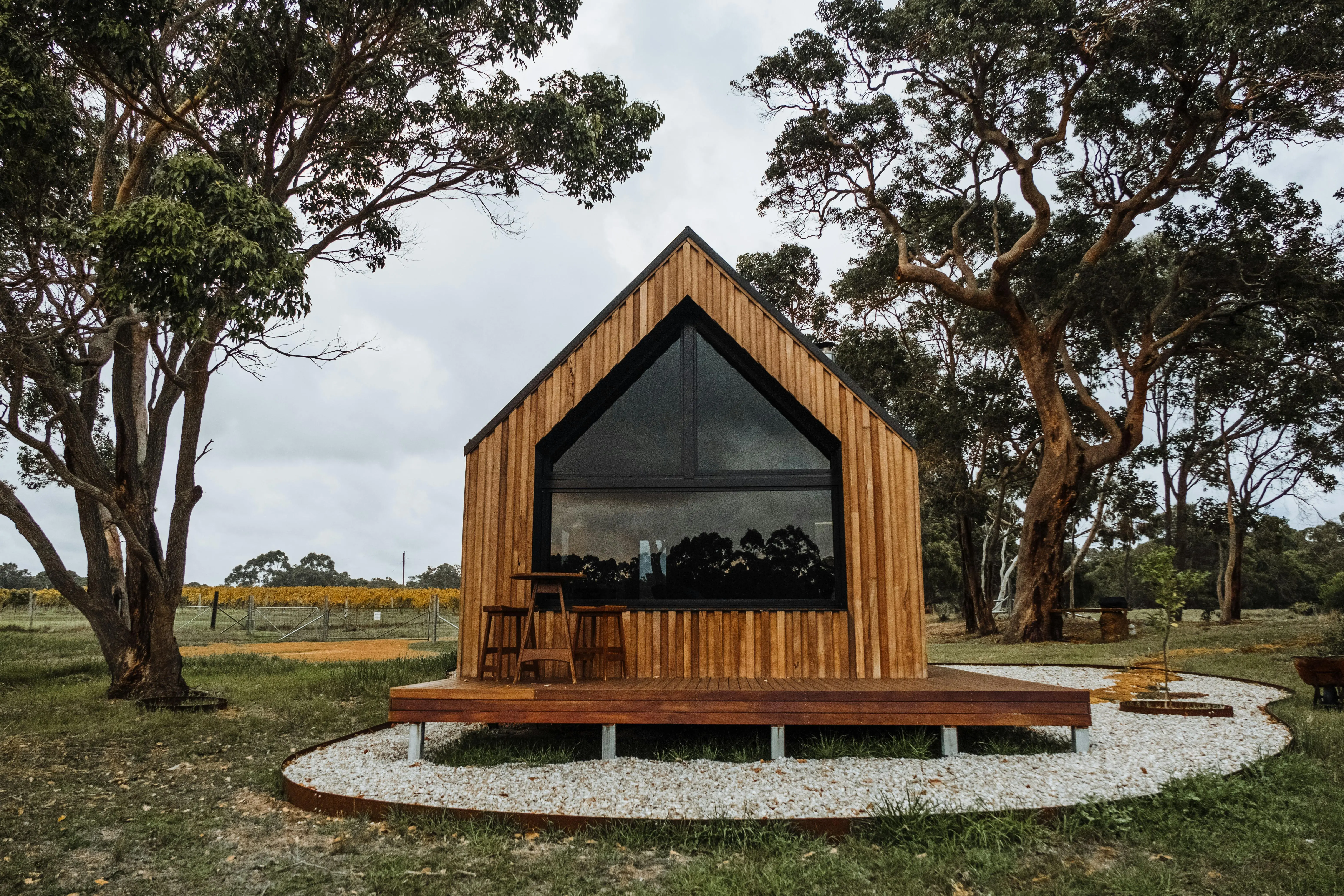 A small wooden A-frame house with a wrap-around deck sits in a patch of gravel next to some trees.