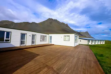 A white house with a wooden deck sits in a grassy field in front of a mountain.
