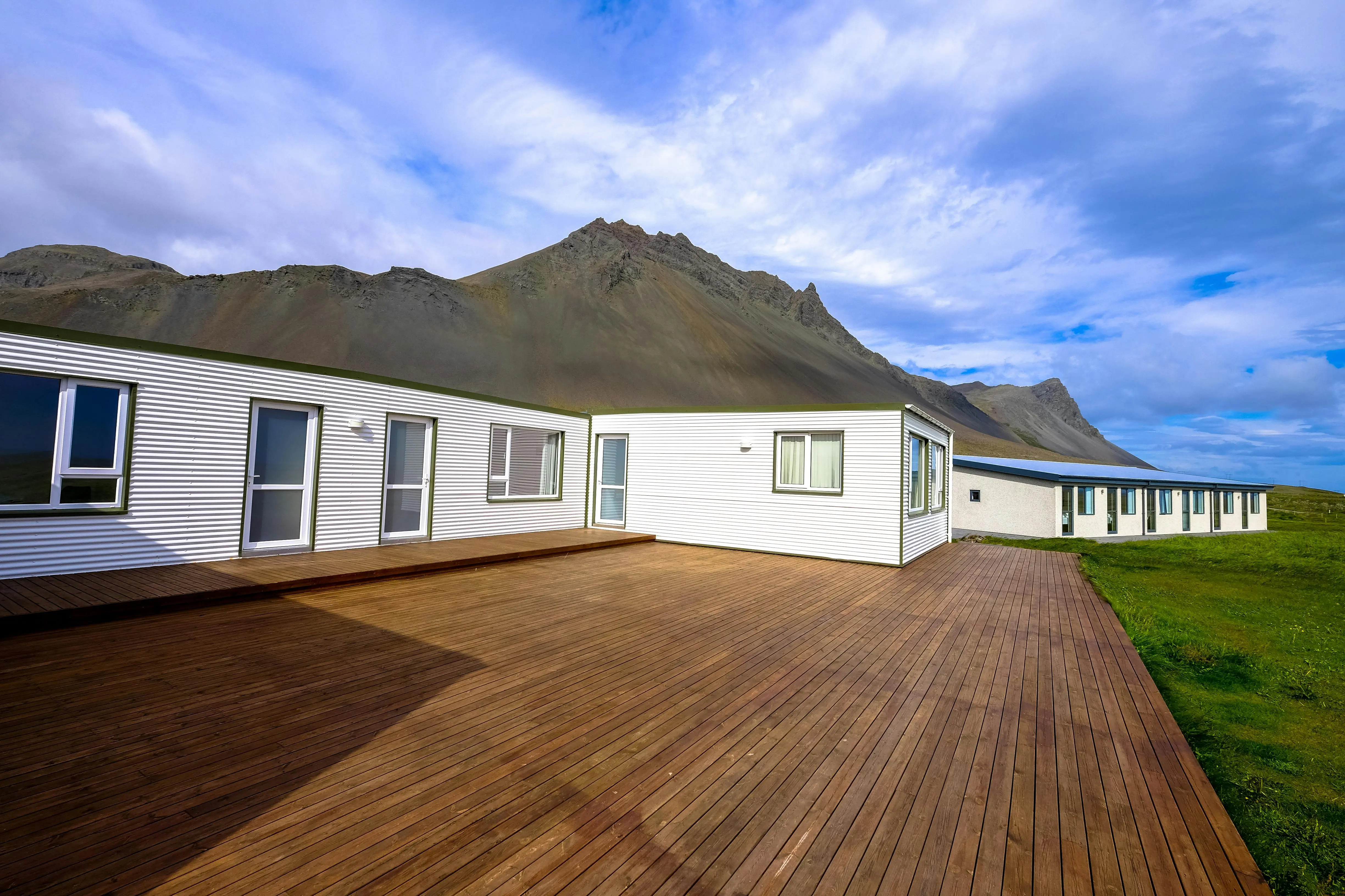 A white house with a wooden deck sits in a grassy field in front of a mountain.