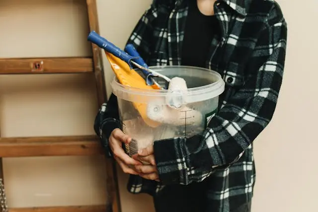 Close-up shot of a person holding a clear bucket filled with painting supplies.