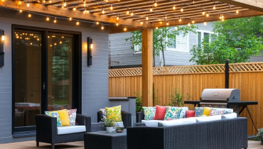 A modern backyard patio with wicker furniture, colorful cushions, a pergola with string lights, a grill, and a wooden fence.