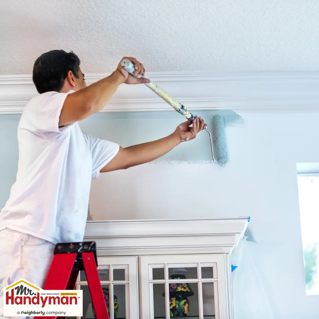 A person on a ladder paints a wall light blue near a ceiling.