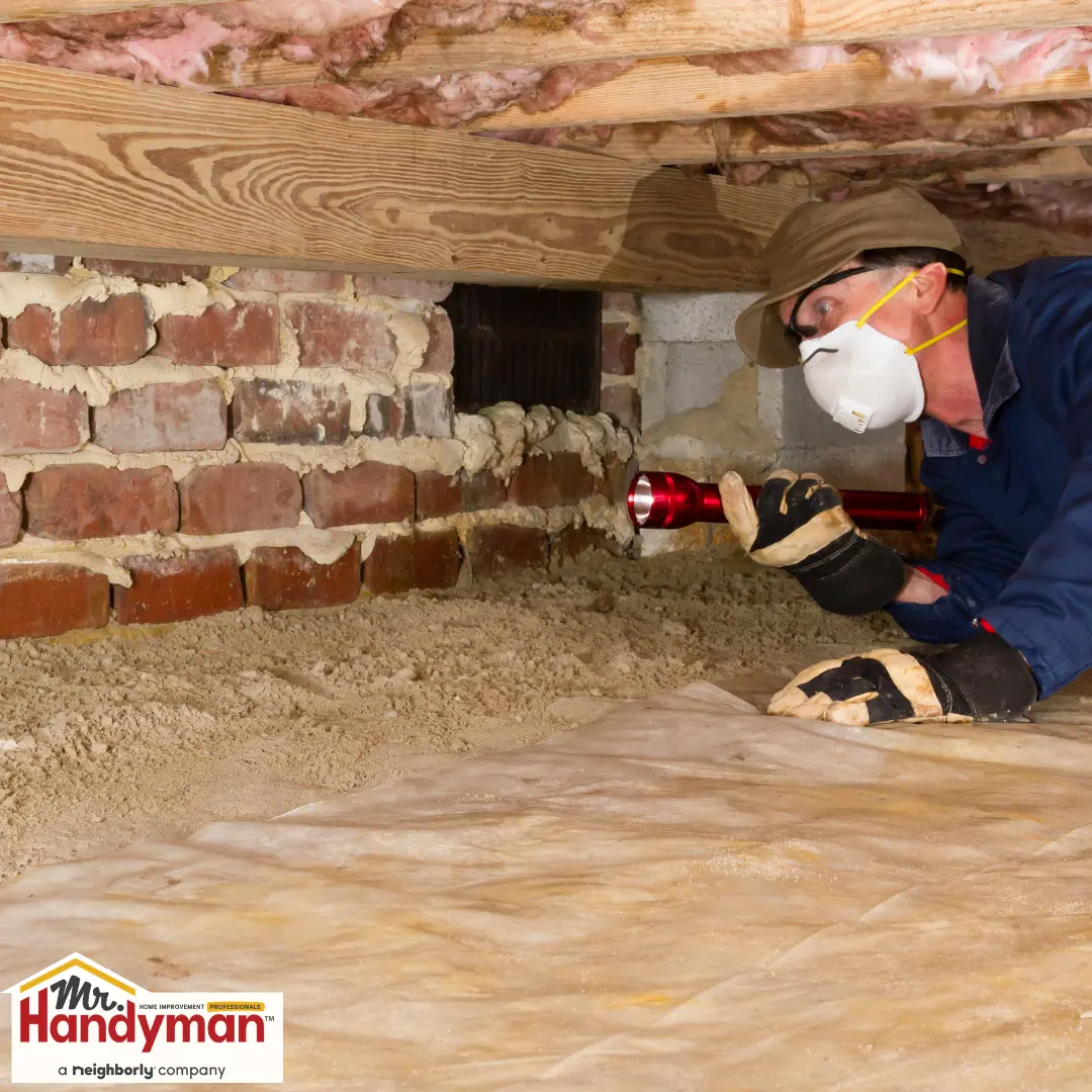 A handyman in protective gear inspects a crawl space.