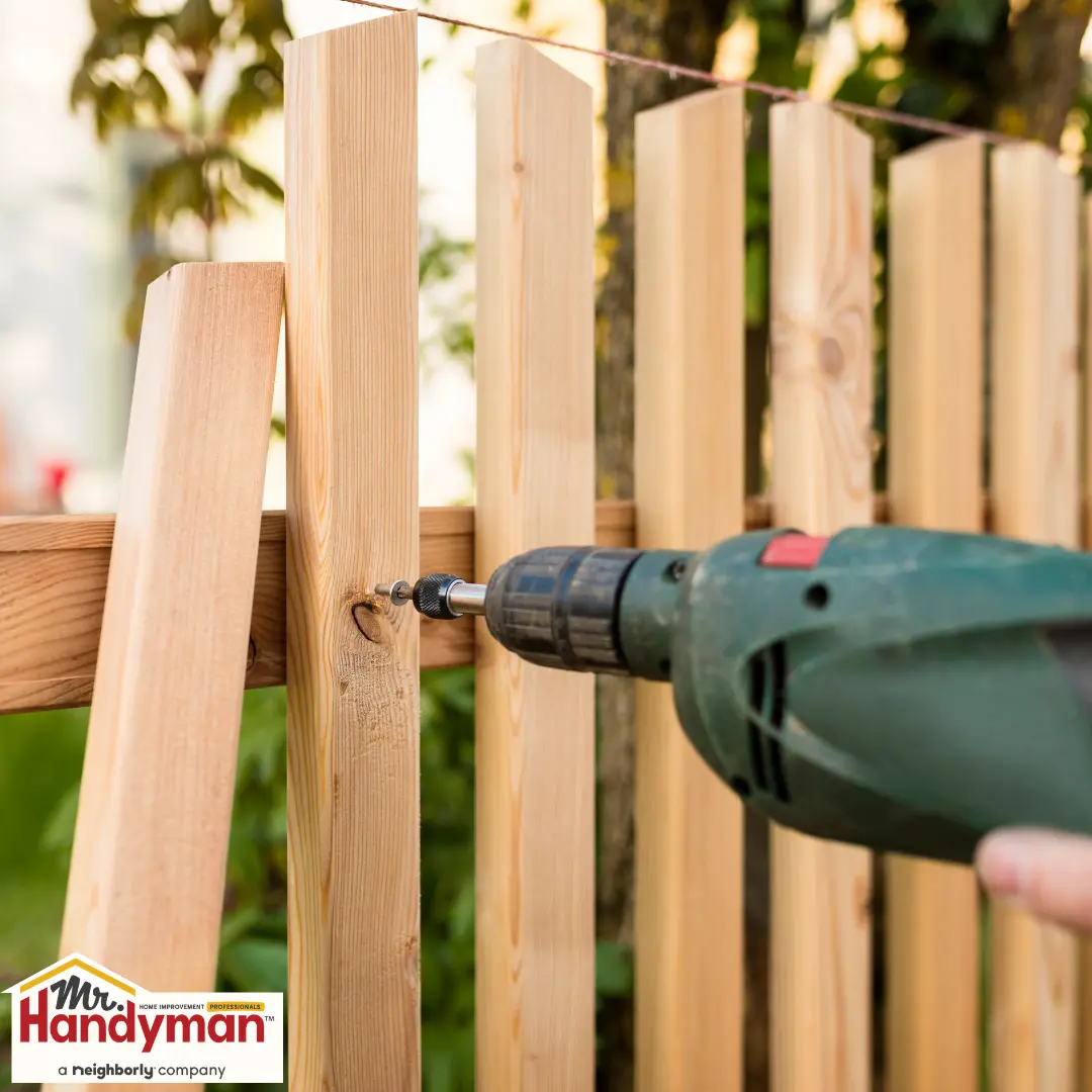 A person using a power drill to fasten a wooden fence post to a horizontal beam.