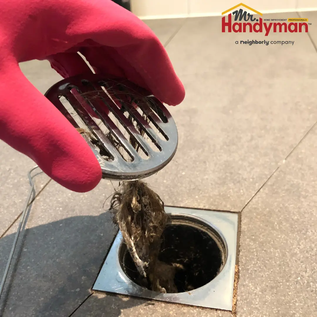 hand in red glove removing a metal drain cover clogged with hair and debris from a floor drain