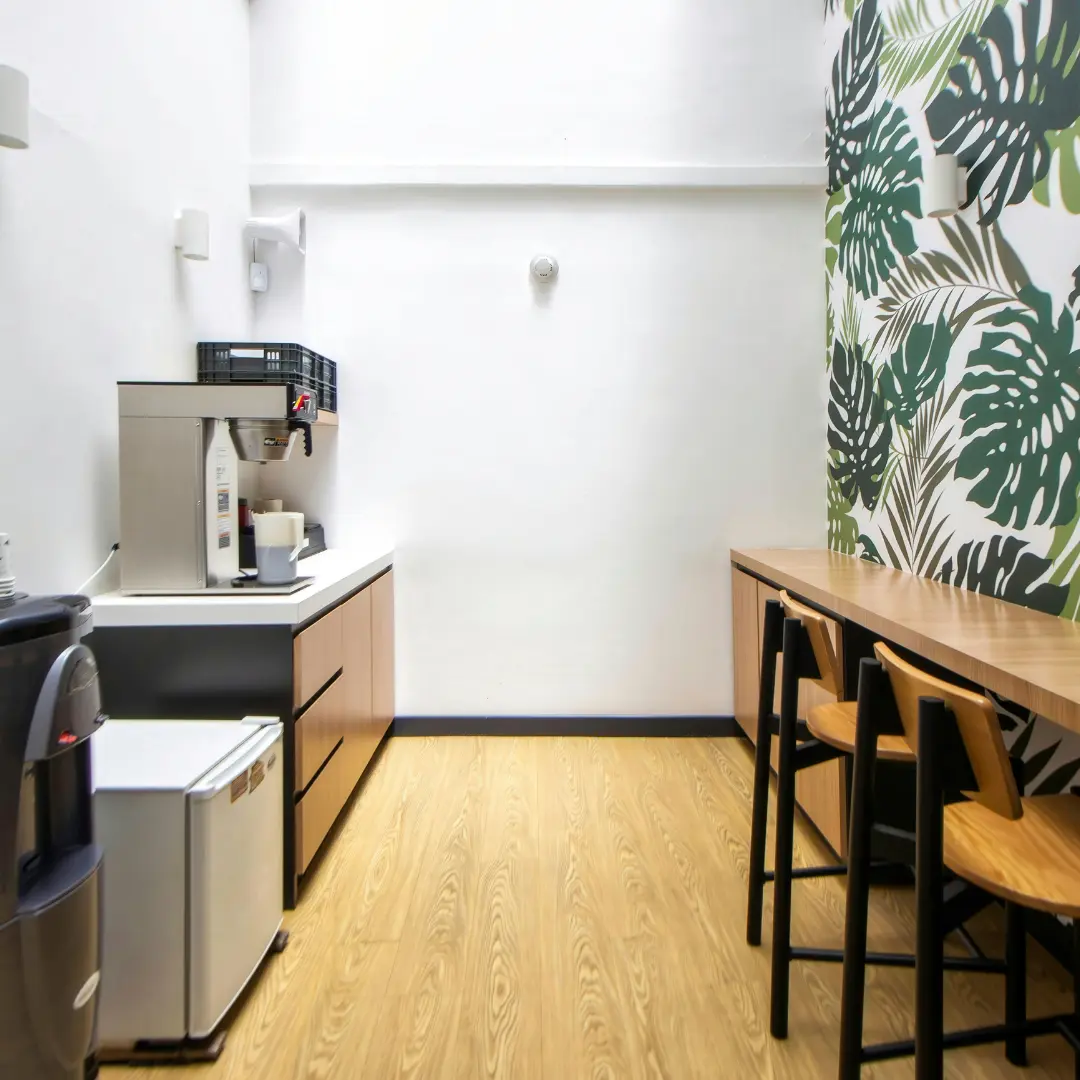 A modern office break room with a coffee station, mini fridge, and a long communal table with high chairs.