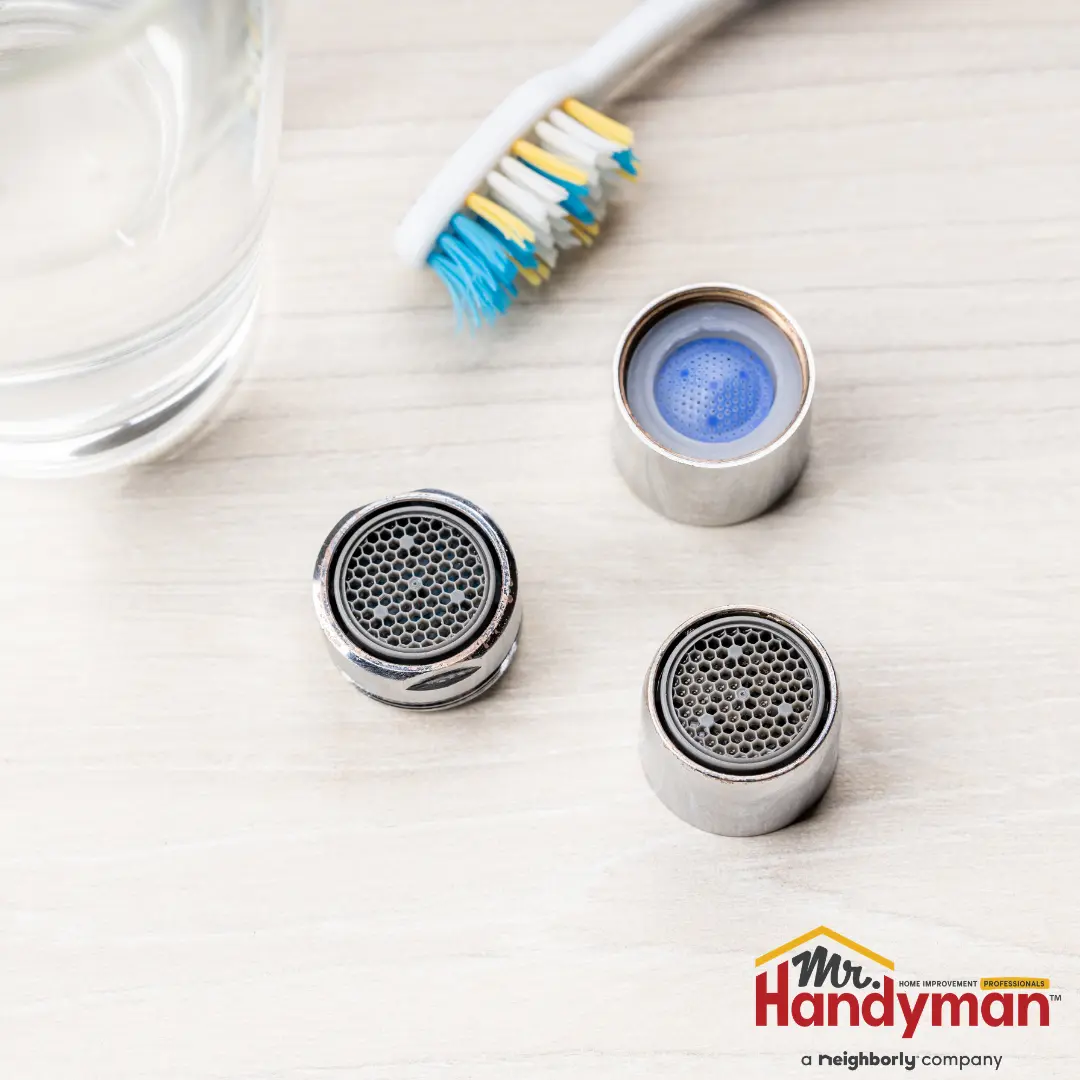A Mr Handyman faucet aerator disassembled into its three main components, placed on a light wooden surface alongside a toothbrush and a glass of water. 