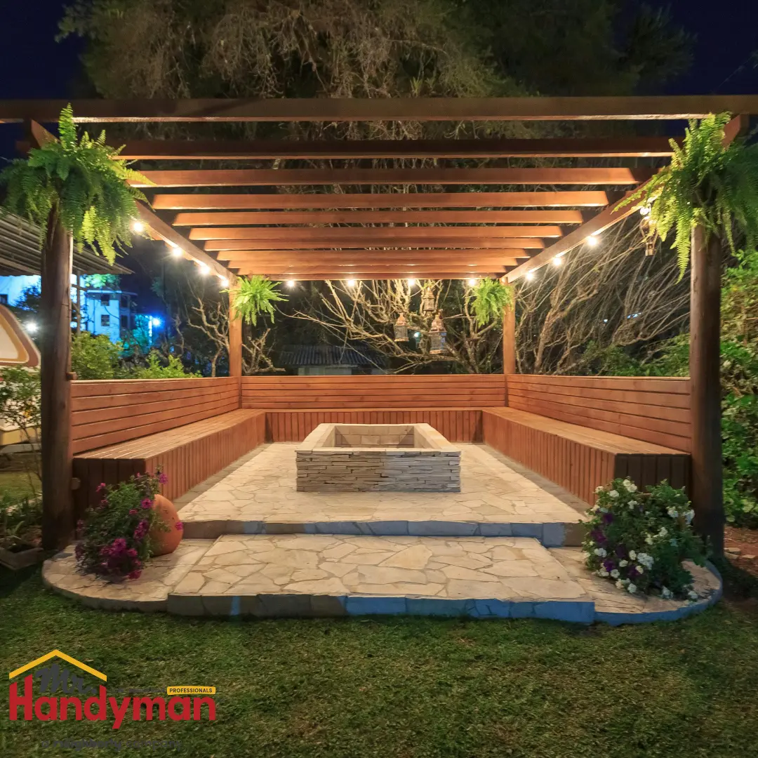 A warmly lit wooden pergola with string lights, surrounded by lush greenery and a stone fire pit at its center.