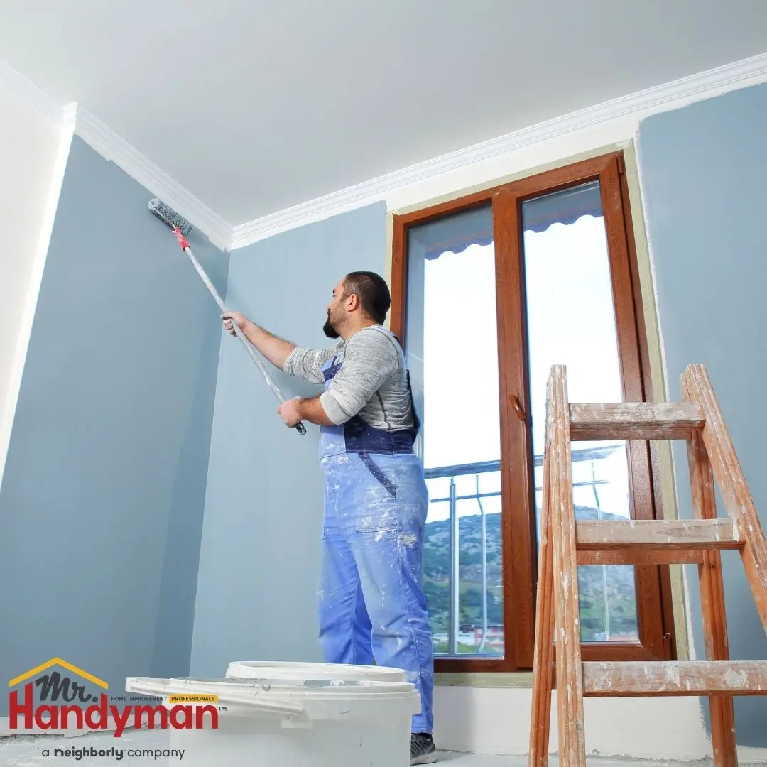 Person painting the upper corner of a room with a roller, standing near a window and a wooden ladder.