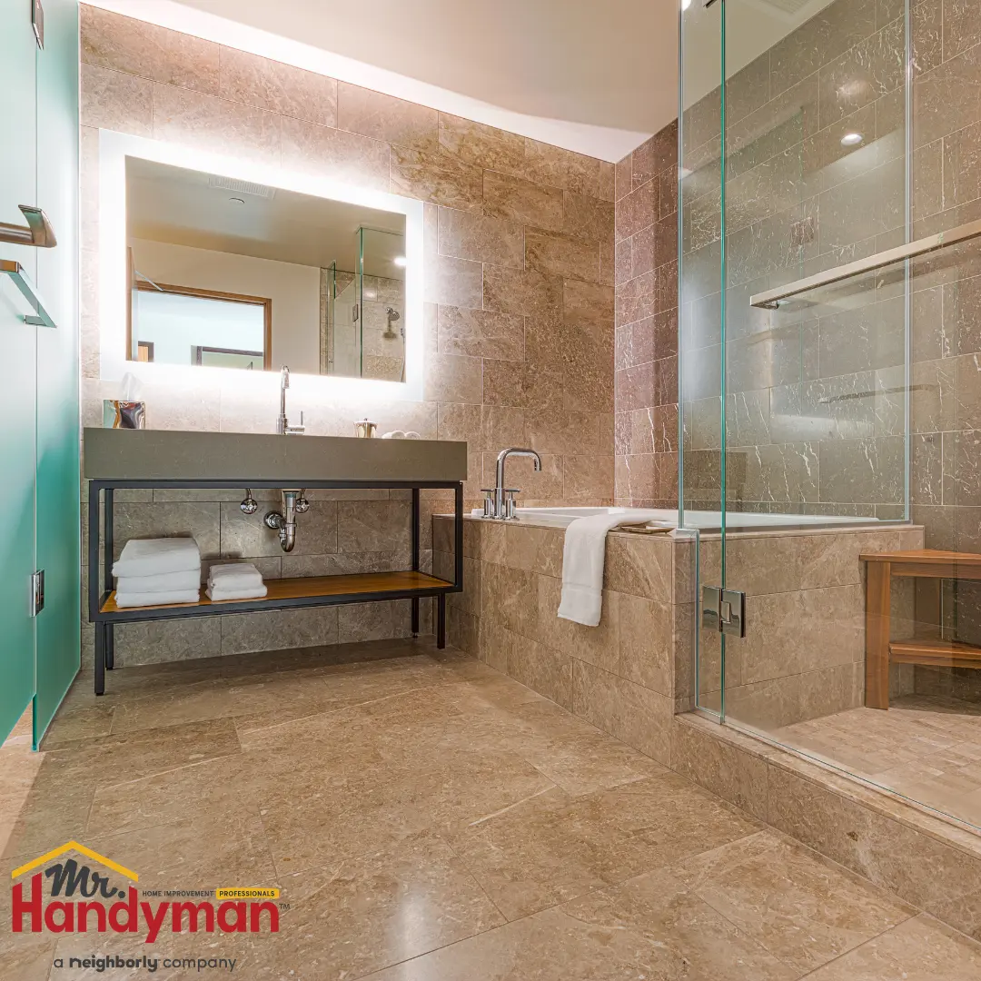 A modern bathroom features tan stone tile walls and floors and a glass-enclosed shower.