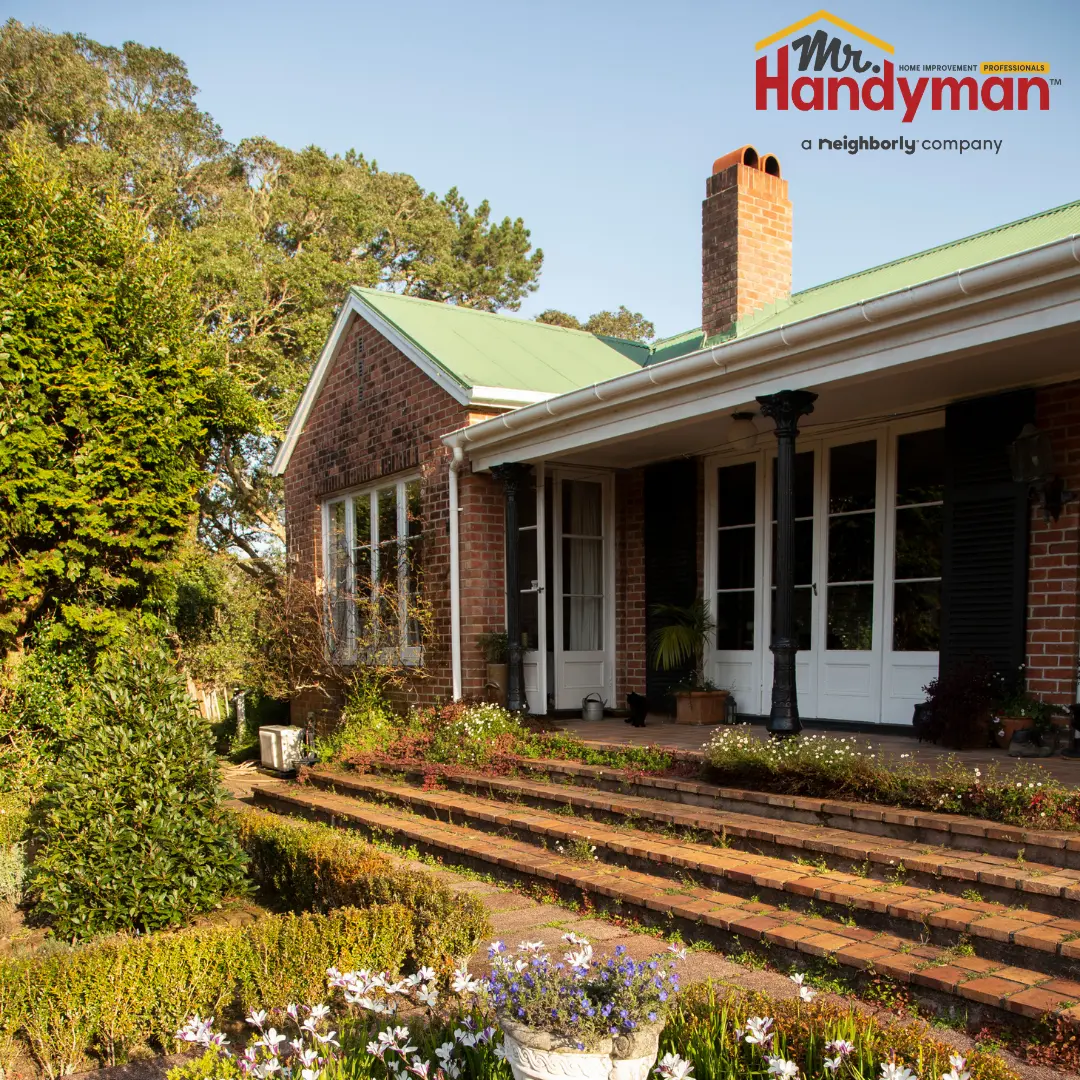 A well-maintained brick house with a green roof, surrounded by a garden featuring colorful flowers and shrubs.