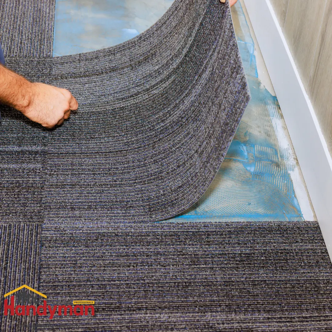 A person laying down a gray striped carpet tile on a blue adhesive surface.