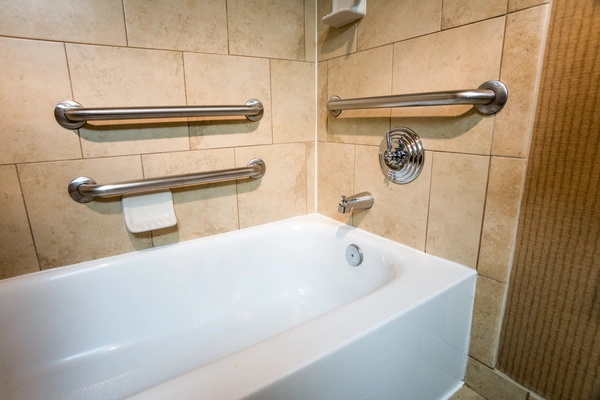 A white bathtub surrounded by beige tiled walls with three stainless steel safety grab bars.