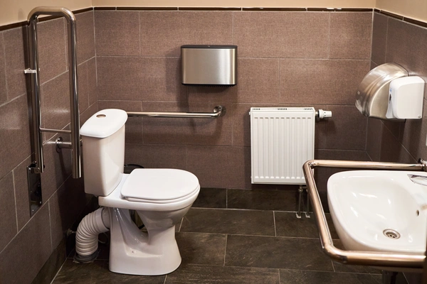 An accessible public restroom featuring a toilet with grab bars, a sink with a protective metal rail, a wall radiator, and a hand dryer.