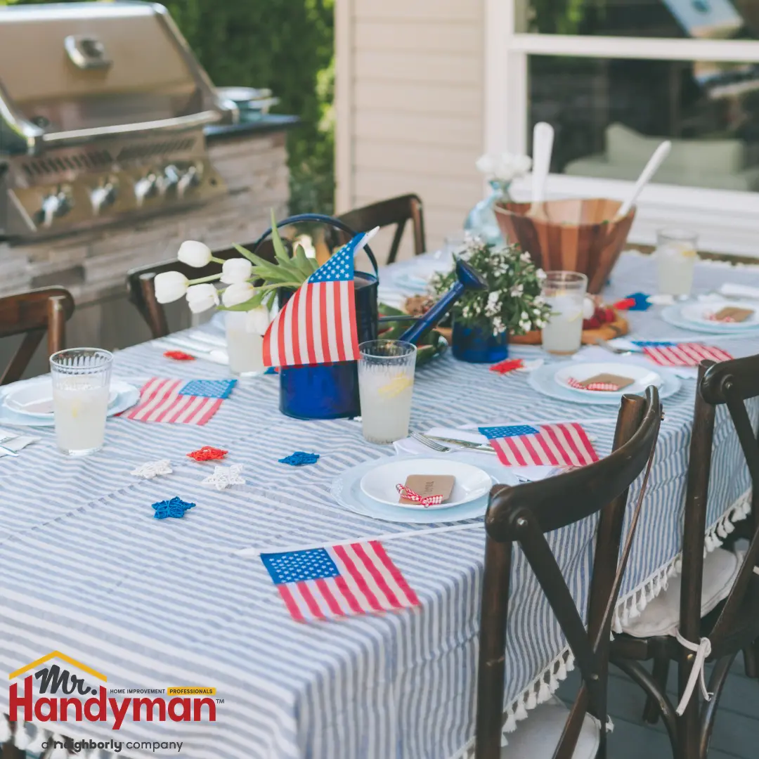An outdoor table with a striped tablecloth, small American flags, lemonade, snacks, and a grill in the background.